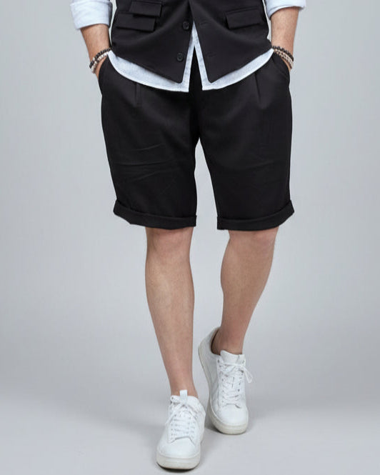 Model in front of light grey backdrop wearing black vest and shorts and white shirt. with tanned legs.