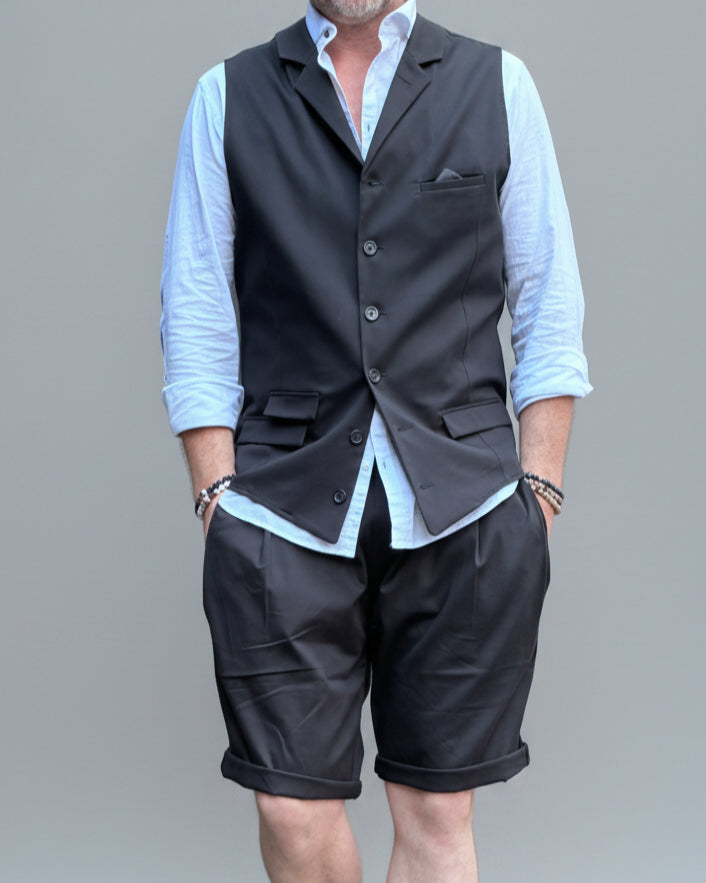 Model in front of light grey backdrop wearing black vest and shorts and white shirt. with tanned legs.