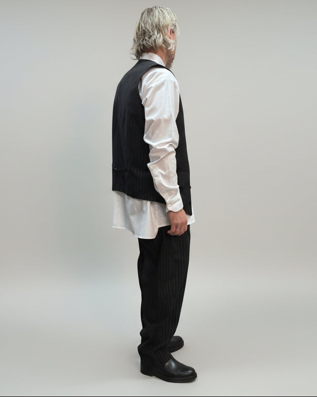 MODEL FACING ON THE SIDE WEARING BLACK PINTRIPE VEST AND LOOSE PANTS IN A RELEXED POSITION