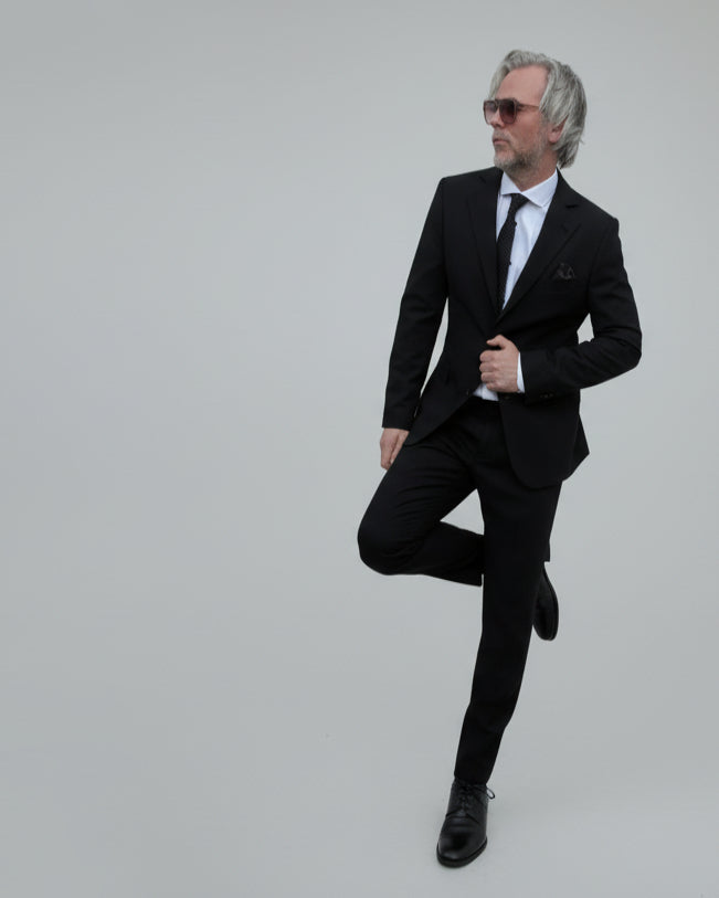 Model leaning against light grey backdrop wearing black suit and white shirt.