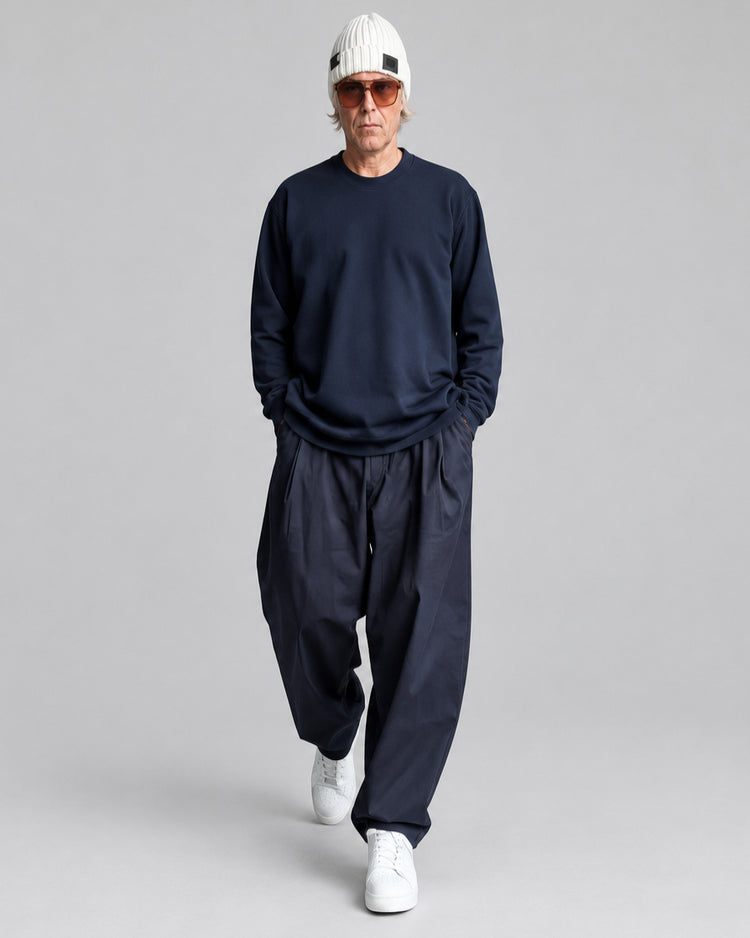 A man wearing a dark navy long-sleeve crewneck sweatshirt, matching dark navy baggy pants, white sneakers, and a white knit beanie with orange-tinted sunglasses, walking in front of a white painted brick wall.
