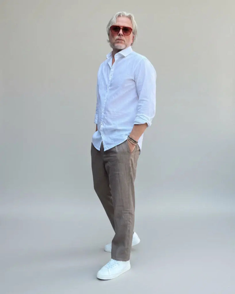 Model standing in front of light grey backdrop wearing brown linen pants and white linen shirt and sneakers
