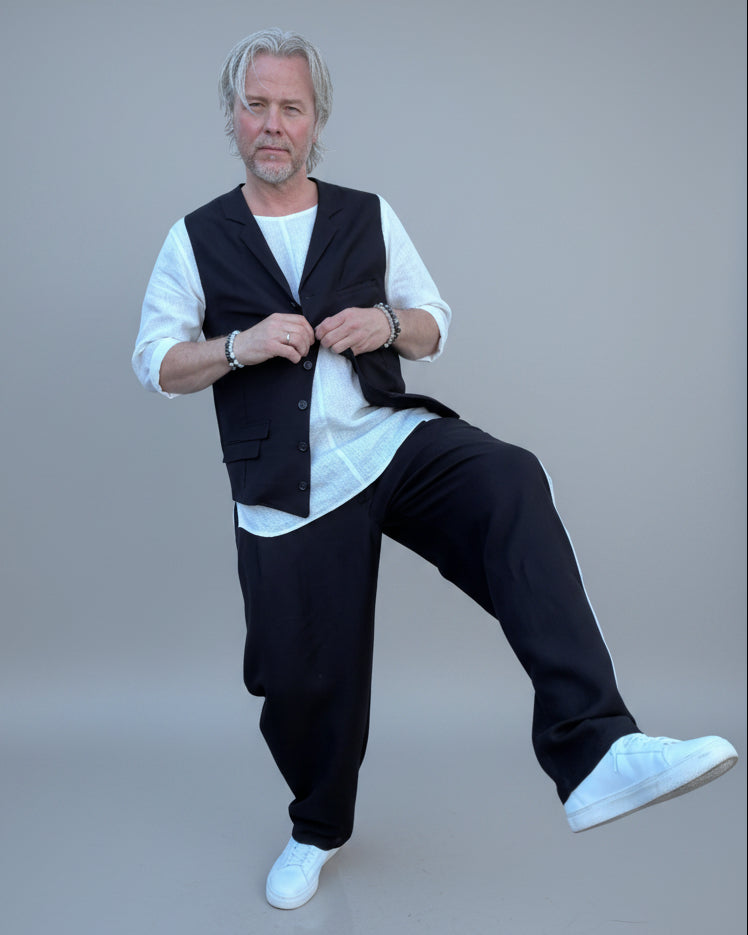 Person wearing a black vest over a white shirt with dark pants and white shoes, in front of light grey backdrop.