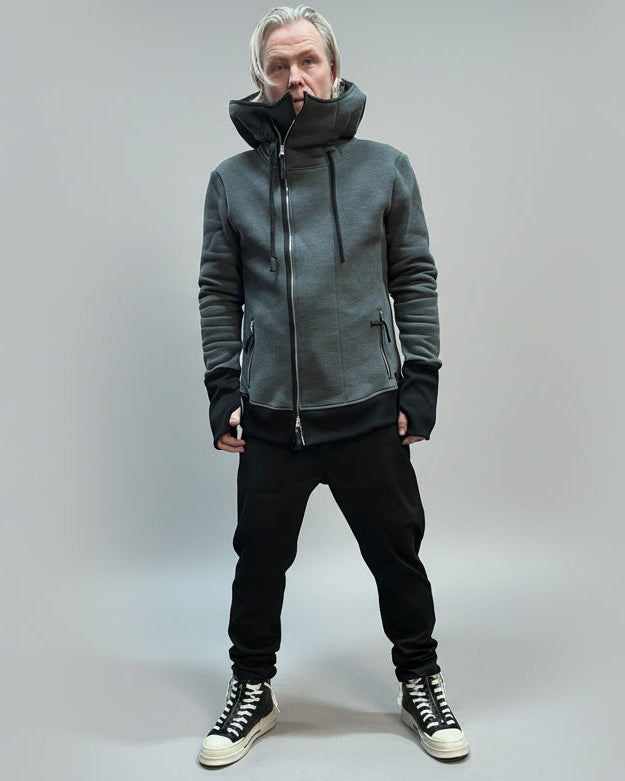 Person wearing a gray hoodie and black pants standing against a light grey curtain.