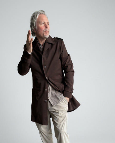 MODEL STANDING IN FRONT OF L. GREY BACKDROP WEARING A BROWN TRENCH COAT