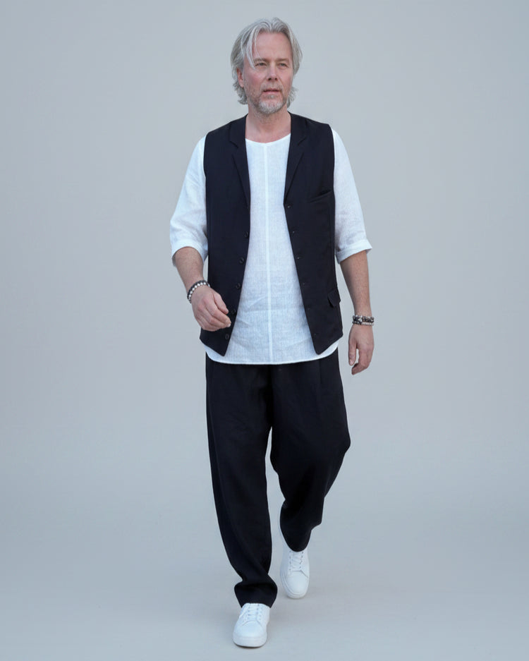 Person wearing a black vest over a white shirt with dark pants and white shoes, in front of light grey backdrop.