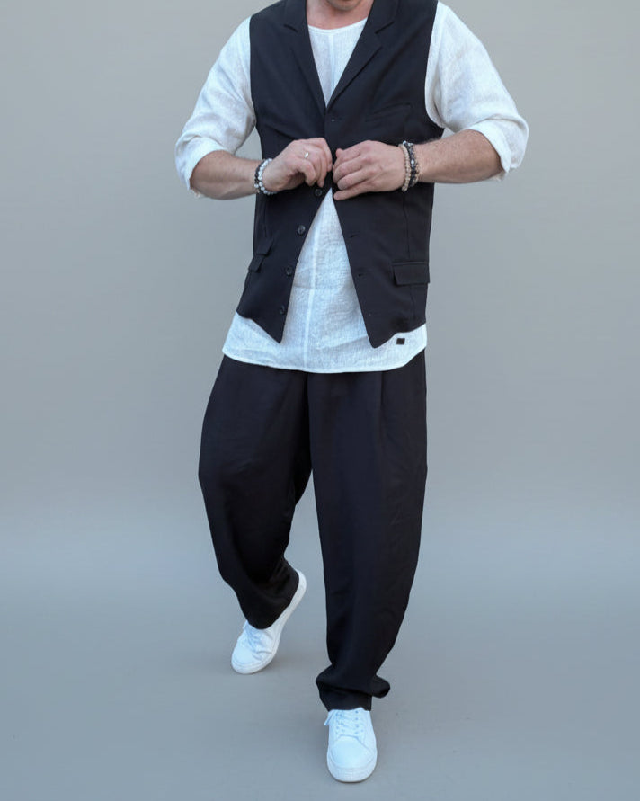 Person wearing a black vest over a white shirt with dark pants and white shoes, in front of light grey backdrop.