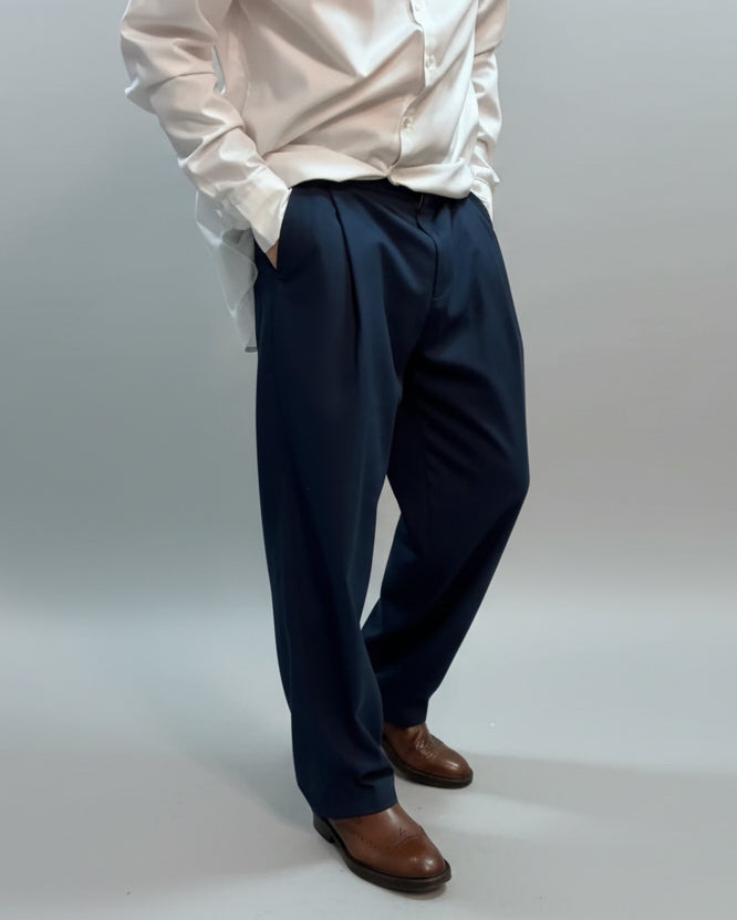 Person wearing a white shirt and navy pants in front of a light grey backdrop with shirt loose and hands in pocket.