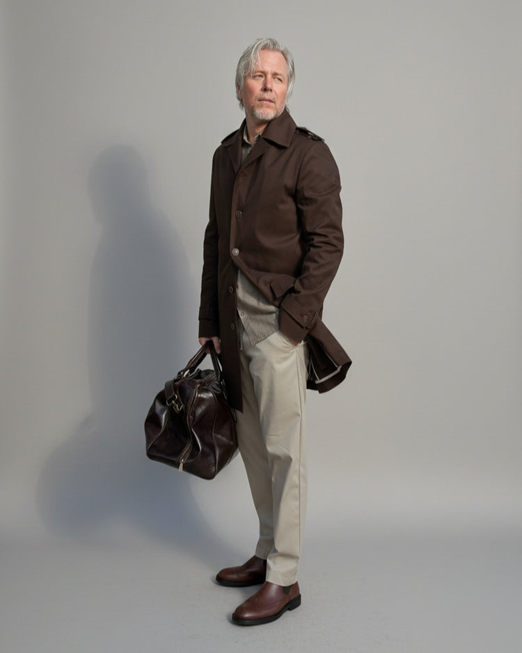 MODEL STANDING IN FRONT OF L. GREY BACKDROP WEARING A BROWN TRENCH COAT HOLDING A BROWN CRUMPY LEATHER BAG.