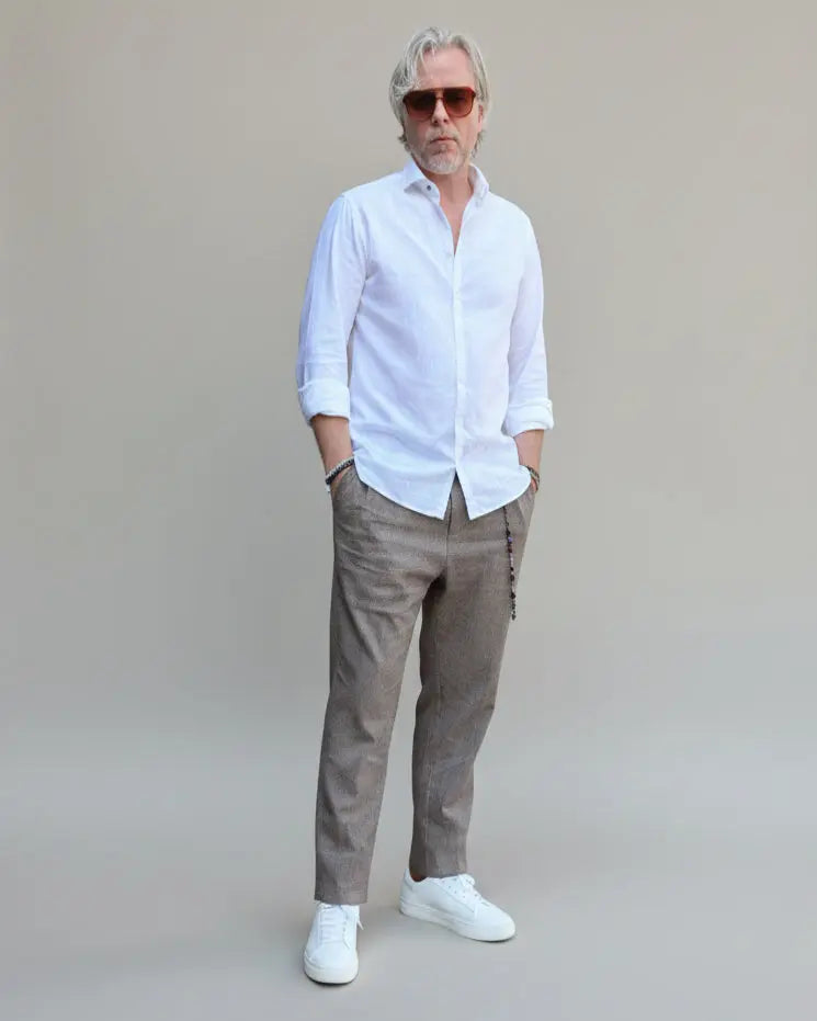 Model standing in front of light grey backdrop wearing brown linen pants and white shirt and sneakers