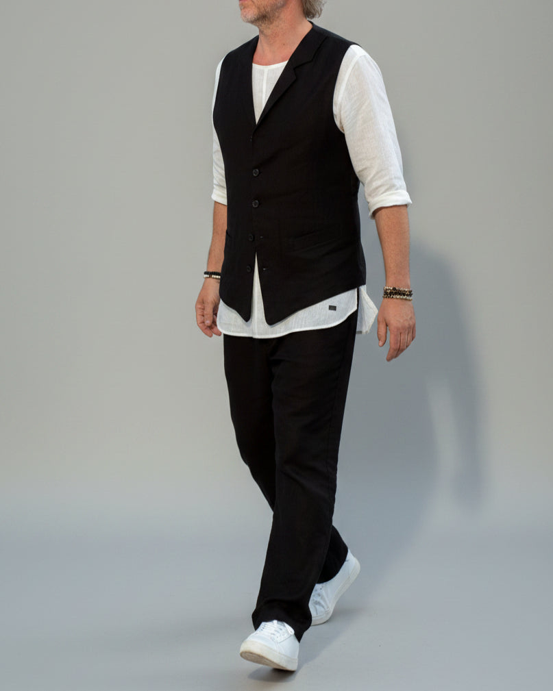 Person wearing a black vest over a white shirt with dark pants and white shoes, in front of light grey backdrop.