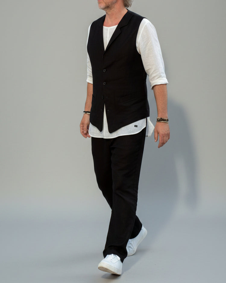 Person wearing a black vest over a white shirt with dark pants and white shoes, in front of light grey backdrop.