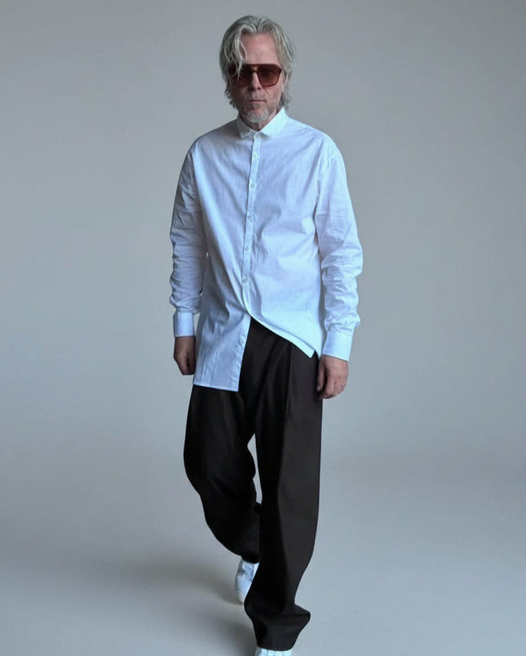 Man wearing a white shirt and sunglasses standing against a white brick wall.