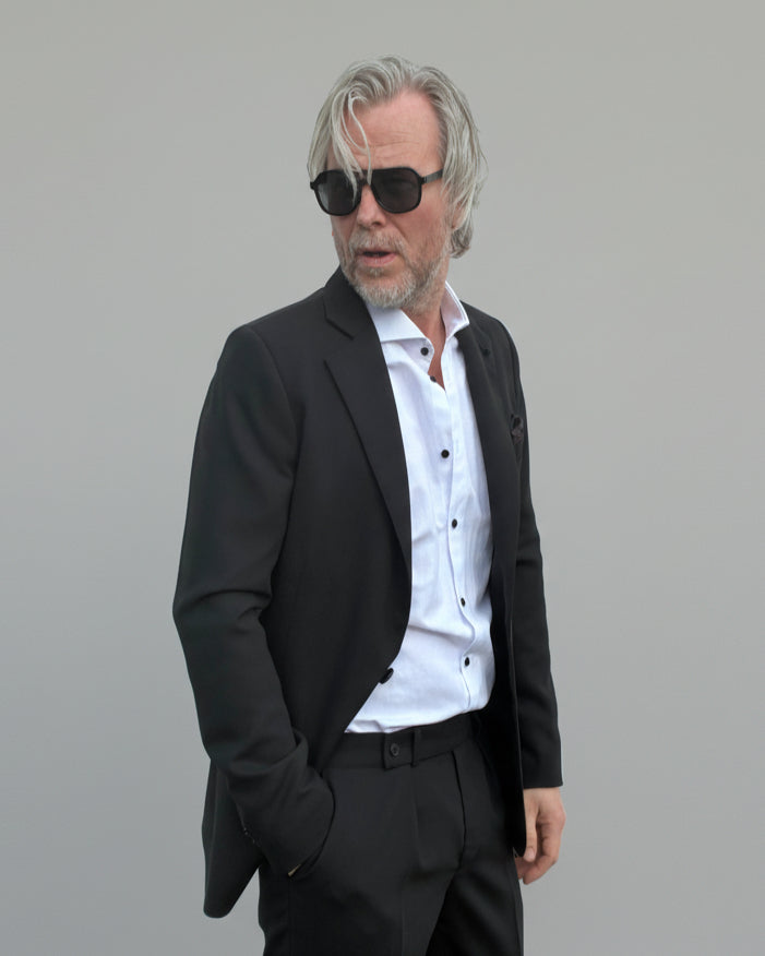 Model standing in front of light grey backdrop wearing black suit and white shirt.