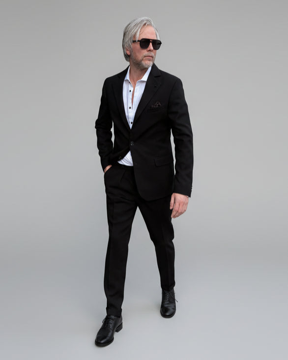 Model standing in front of light grey backdrop wearing black suit and white shirt.