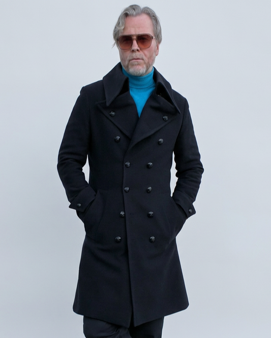 A man wearing a blue double-breasted coat with black metal buttons over a bright blue turtleneck, dark trousers, and brown leather shoes, standing against a light blue wall.