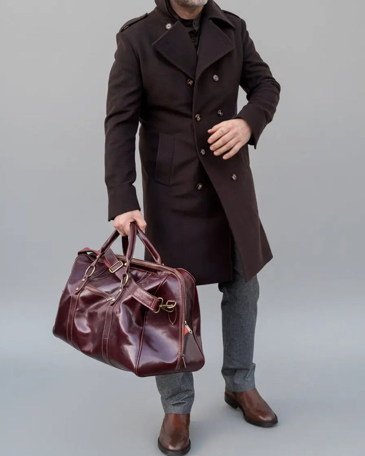 Man in a dark coat holding a brown leather bag against a white brick wall. Head is cut off from neck up.
