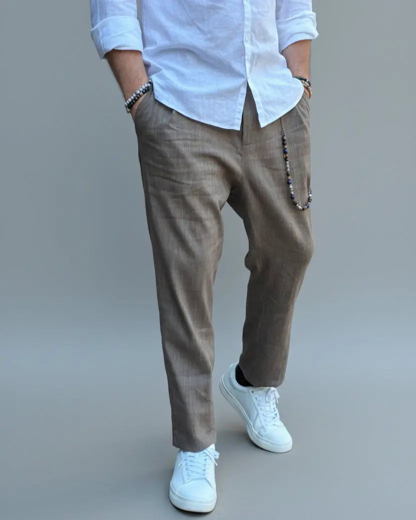 Model standing in front of light grey backdrop wearing brown linen pants and white shirt and sneakers