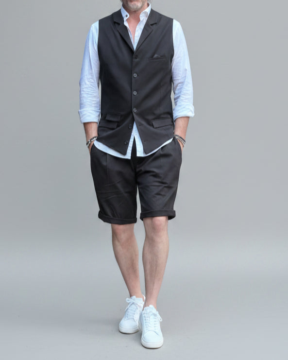Model in front of light grey backdrop wearing black vest and shorts and white shirt. with tanned legs.