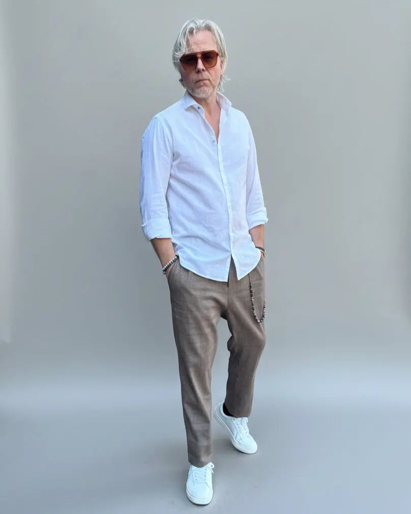 Model standing in front of light grey backdrop wearing brown linen pants and white shirt and sneakers
