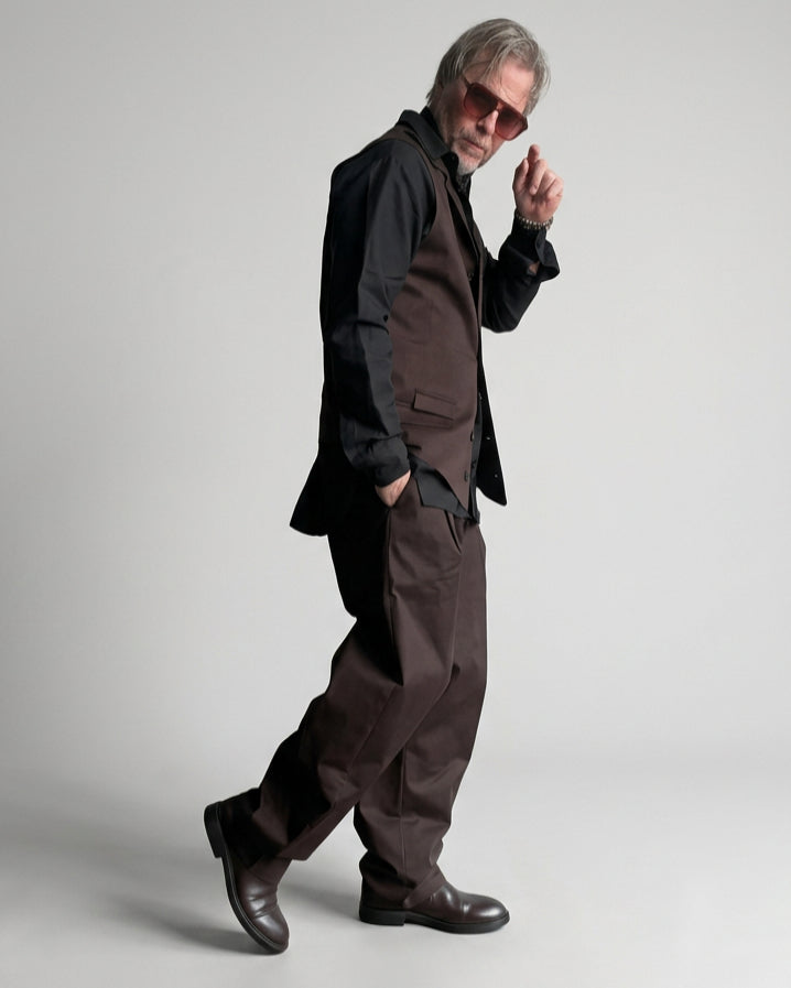 A man wearing a dark brown vest and balloon trousers, black shirt, and sunglasses stands against a white painted brick wall.