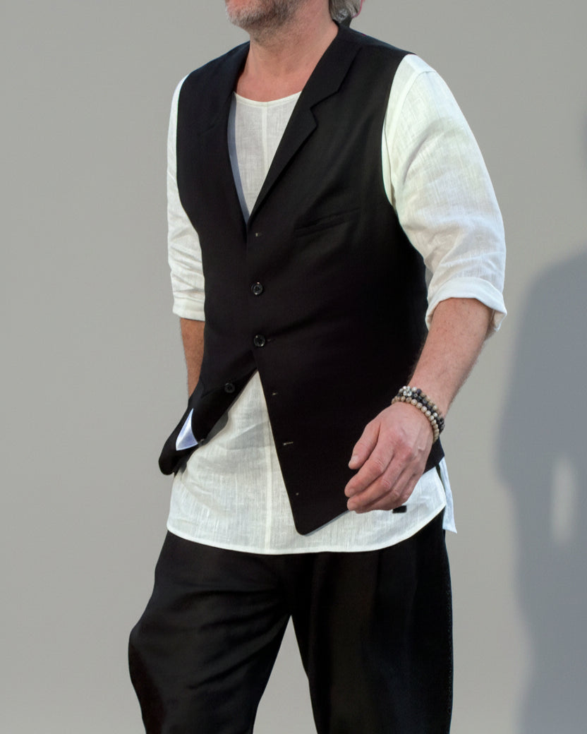 Person wearing a black vest over a white shirt with dark pants and white shoes, in front of light grey backdrop.