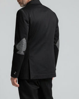 Person standing on a wooden platform with a plain background, wearing a black jacket with spade and diamond elbow patches 