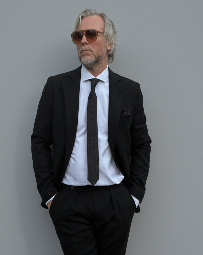 Model standing in front of light grey backdrop wearing black suit and white shirt.