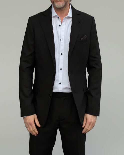Model standing in front of light grey backdrop wearing black suit and white shirt.
