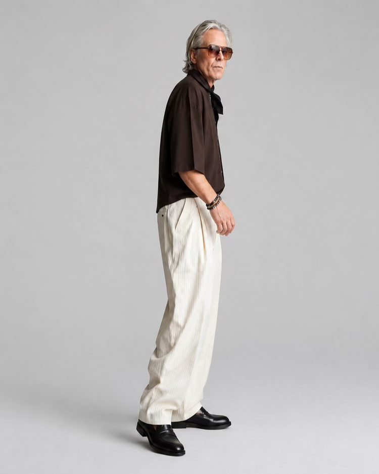 A man wearing a dark brown short-sleeve T-shirt and a silk scarf, cream-colored pleated trousers, and sunglasses stands in profile against a white brick wall.
