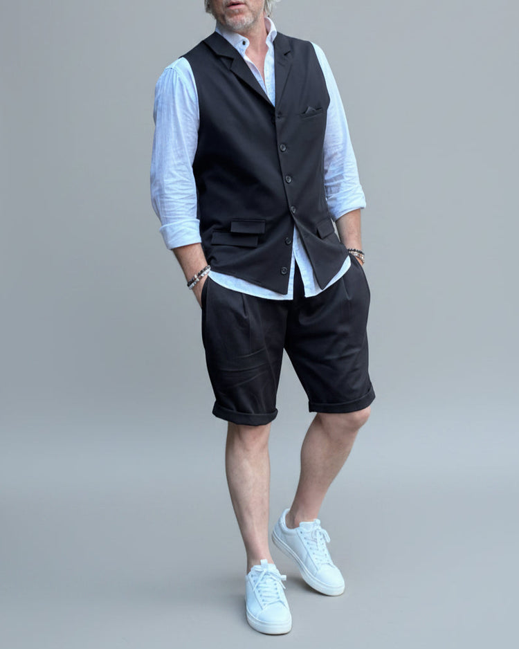 Model in front of light grey backdrop wearing black vest and shorts and white shirt. with tanned legs.