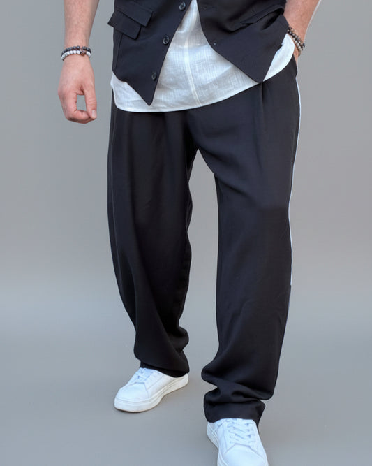 Person wearing a black vest over a white shirt with dark pants and white shoes, in front of light grey backdrop.