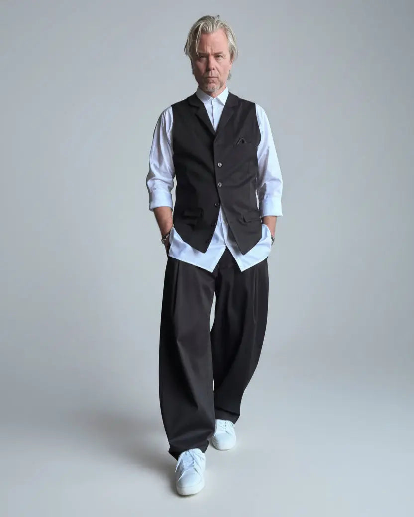 Model in baggy black pants, white shirt and black waistcoat standing in front of light grey backdrop