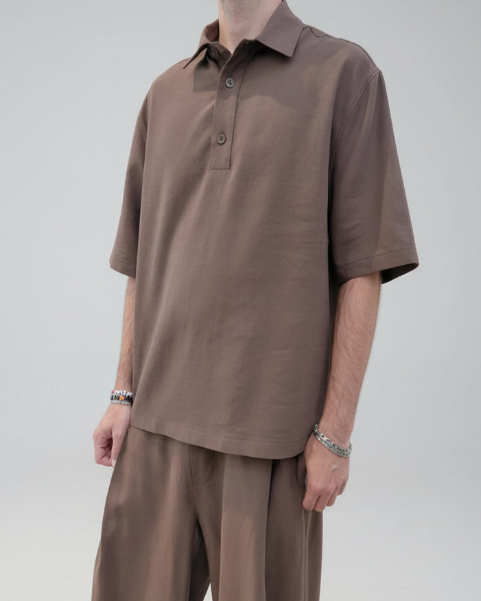 A man wearing a brown short-sleeve polo shirt and matching brown shorts, standing against a plain light gray wall.