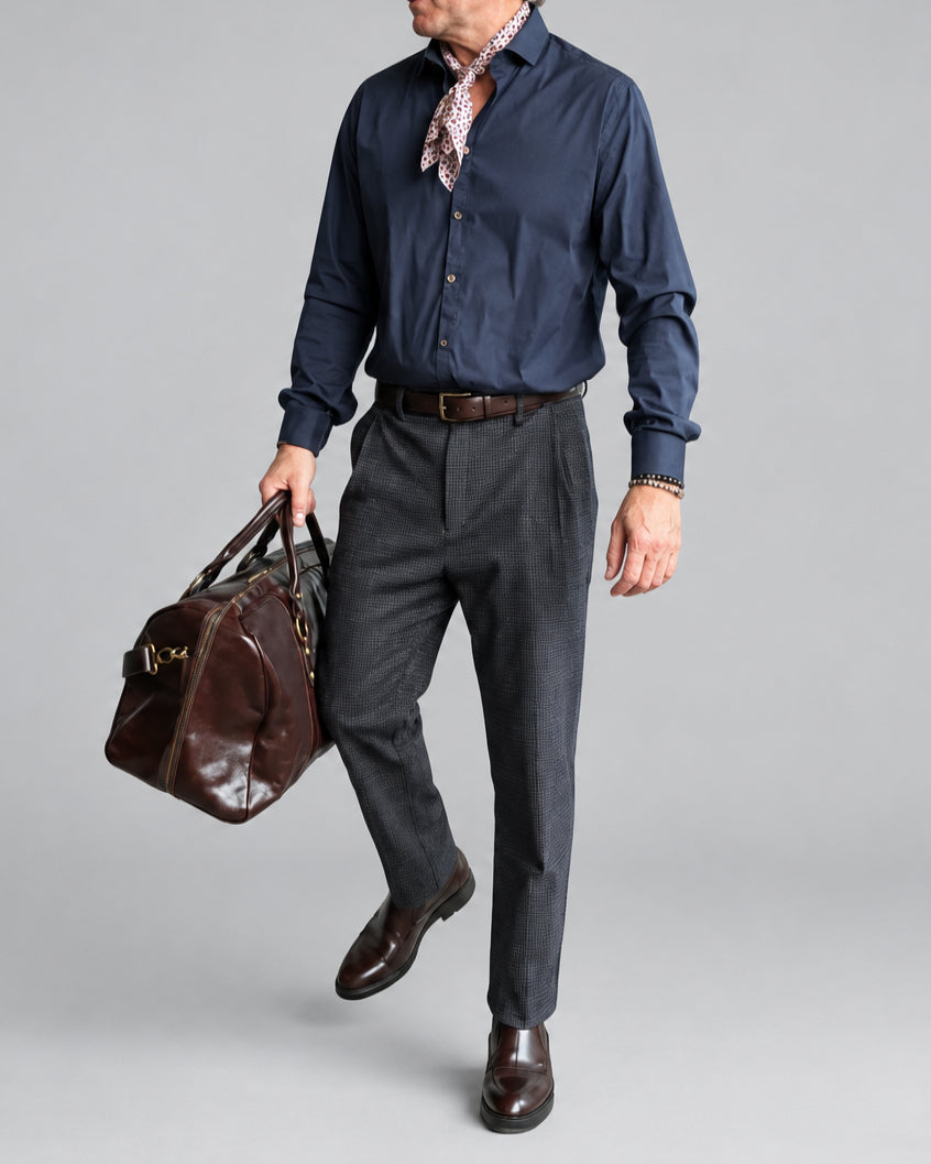 A man wearing a navy blue button-down shirt, blue/gray micro checked trousers, a brown leather belt, and brown leather shoes, carrying a brown leather duffel bag against a white brick wall background.