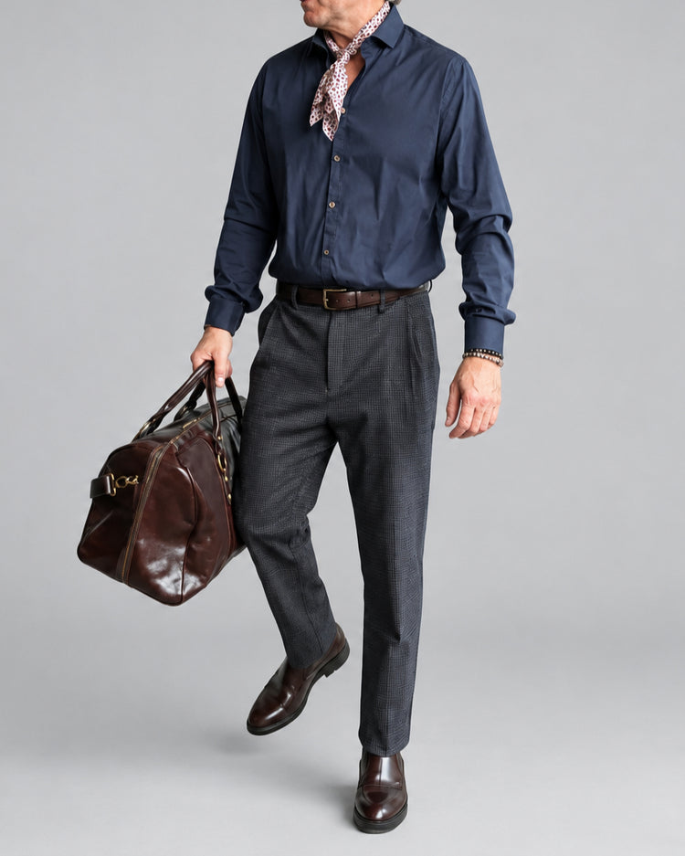 A man wearing a navy blue button-down shirt, blue/gray micro checked trousers, a brown leather belt, and brown leather shoes, carrying a brown leather duffel bag against a white brick wall background.