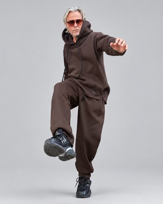 A man with gray hair and sunglasses poses in a brown hooded sweatshirt and matching sweatpants WITH NO CARGO POCKETS ON ITHEM in front of a dark metal door and white brick walls.