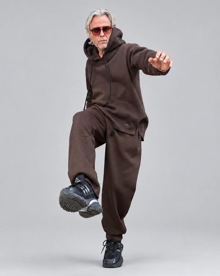 A man with gray hair and sunglasses poses in a brown hooded sweatshirt and matching sweatpants WITH NO CARGO POCKETS ON ITHEM in front of a dark metal door and white brick walls.
