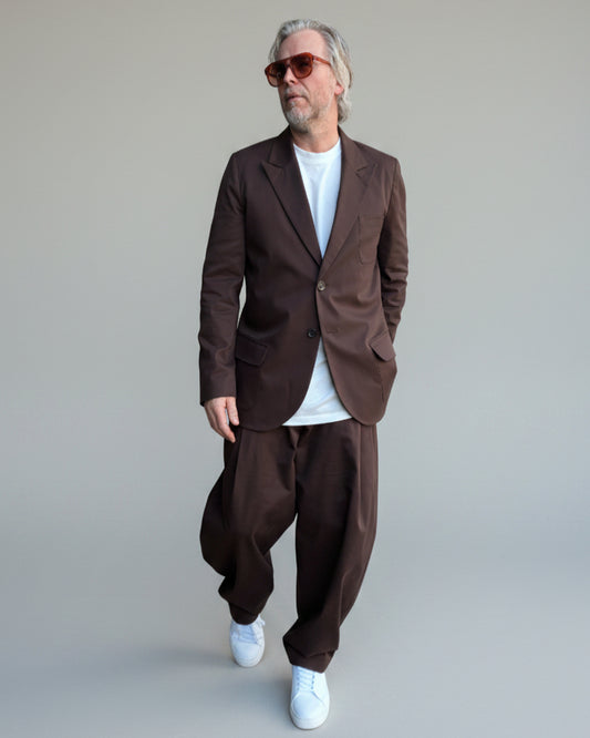 Model wearing brown oversized suit with white shoes and T-shirt in front of light grey backdrop.