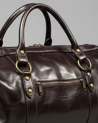 Brown leather duffel bag with a shoulder strap and 1 puller on the side pocket on a textured light grey surface