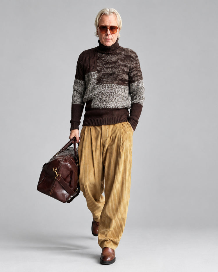 A man walking outdoors against a white brick wall, wearing a patchwork brown and gray patterned turtleneck sweater, loose mustard yellow corduroy pants, brown shoes, and sunglasses, while carrying a dark brown leather duffel bag.