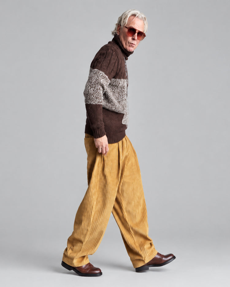 A man wearing a brown and beige patchwork roll-neck sweater, mustard yellow corduroy trousers, and brown leather shoes stands in profile against a white brick wall.